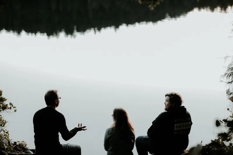 Silhouettes of three people sat talking in front of a lake