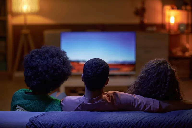 Photograph of three people sitting on a sofa watching the television, with atmospheric lighting and backs facing the camera.