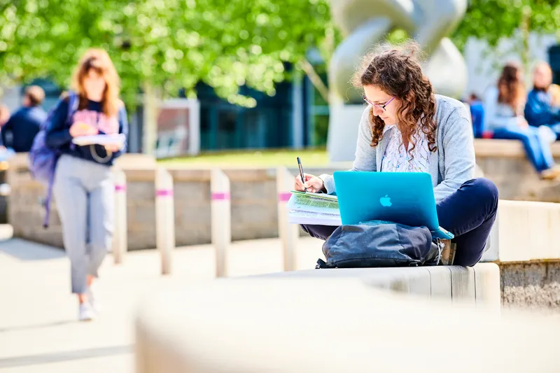 A student sitting outside on campus with a laptop and textbooks