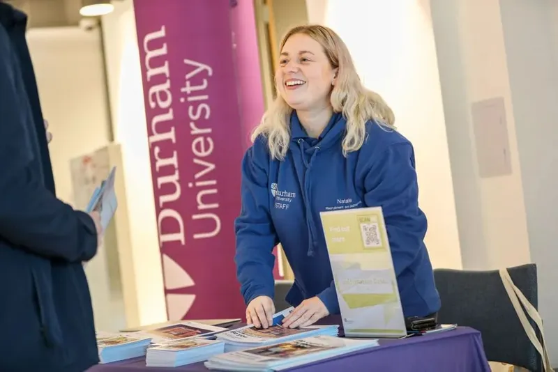 Person smiling whilst handing out Durham University leaflets from behind a desk