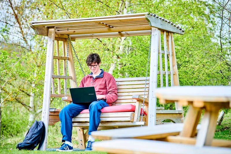 Male student sits on a wooden garden bench swing as he uses his laptop