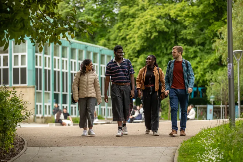 Four students walk towards the viewer. They are chatting as they walk. In the background are a mix of buildings include a red brick building and zinc green building and lots of trees (including a willow tree) in full leaf and grassy banks. It’s an overcast day.
