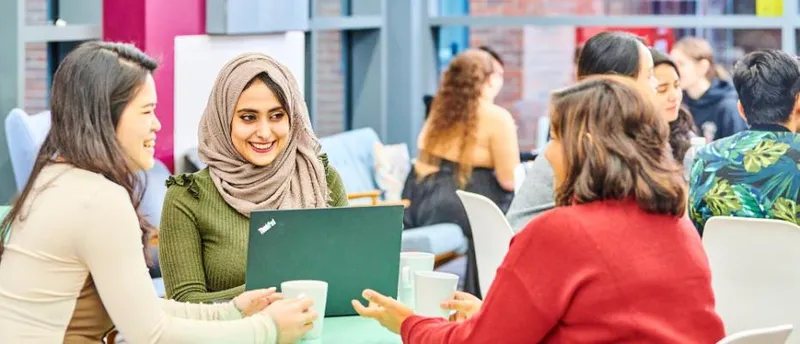 Group of postgraduate students smiling around a laptop with mugs