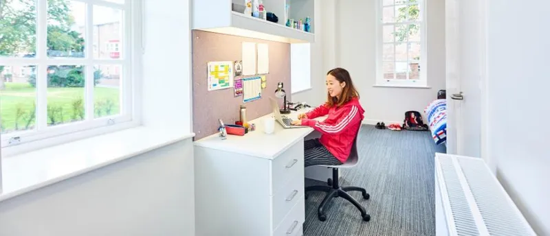 Postgraduate student wearing red smiling at a laptop in their bedroom at Ustinov College