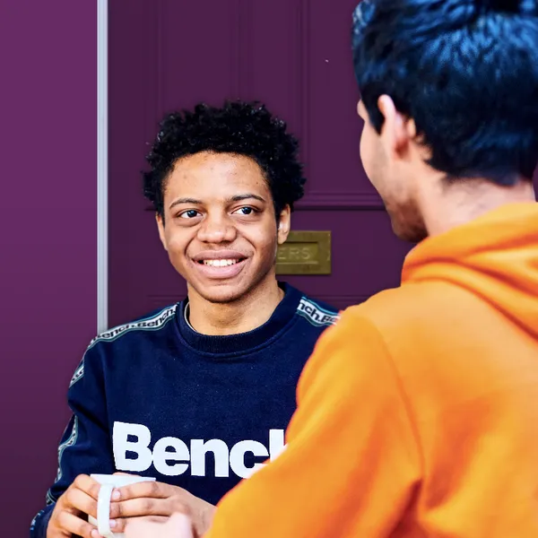 A male student with a hot drink talking to another person facing away from the camera, in front of a purple house door