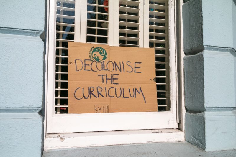 Handwritten 'Decolonise The Curriculum' protest sign on brown cardboard box, mounted in window with green ARTH SERIES logo