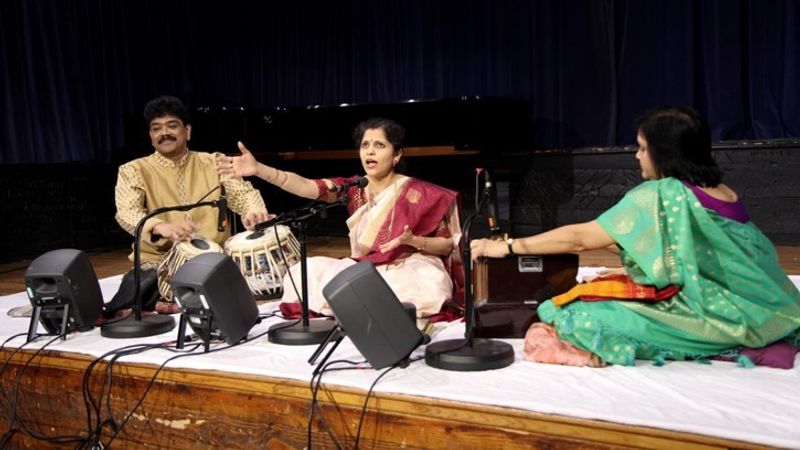 A photograph of a group of musicians sitting on the floor playing instruments, the woman in the centre of the picture is singing
