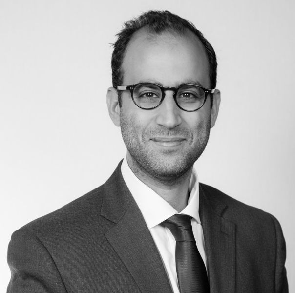 Black and white photo of ABDEL HAKAM wearing a suit, black glasses. He has short brown hair and is smiling.