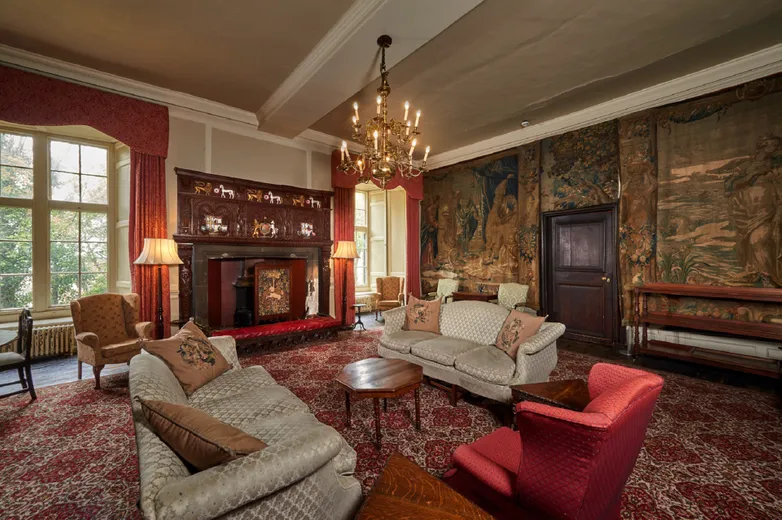 A coloured photograph showing University College Senior Common Room. At the centre of the room is an ornate fireplace, and along the walls are hung tapestries. There is furniture at the edge and at the centre of the room.