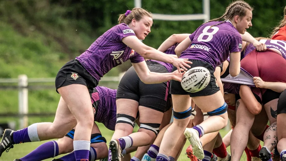 Two teams of female rugby players wearing purple tops, and red and white tops, playing against each other