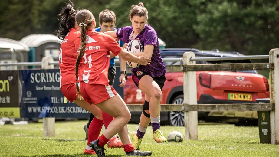 Two teams of female rugby players wearing purple tops, and red and white tops, playing against each other
