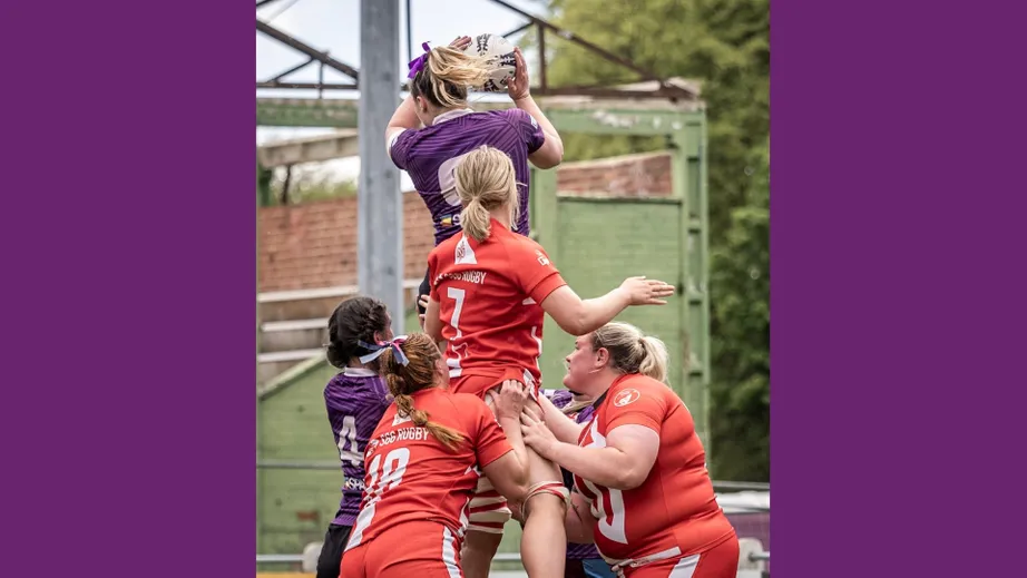 Two teams of female rugby players wearing purple tops, and red and white tops, playing against each other