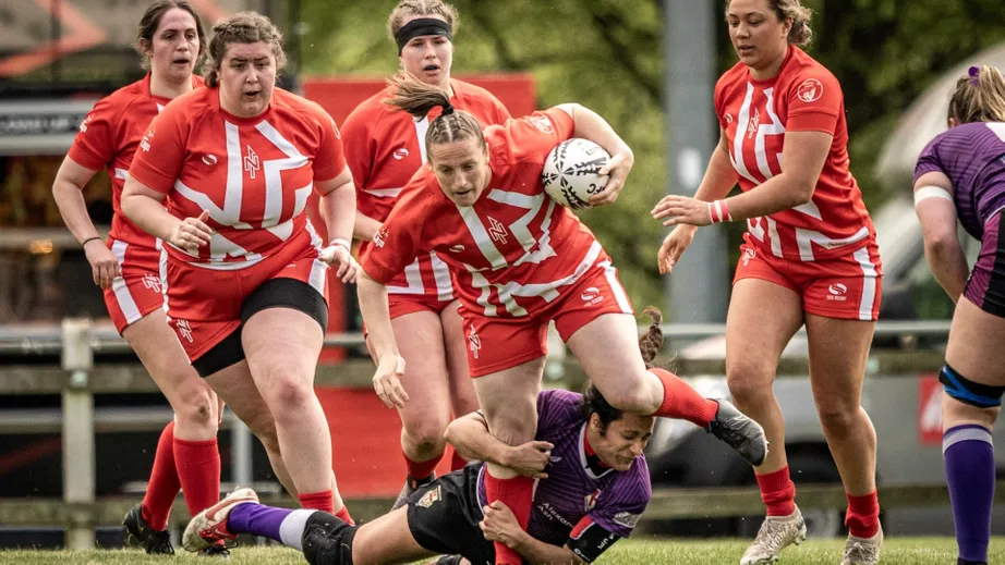 Two teams of female rugby players wearing purple tops, and red and white tops, playing against each other