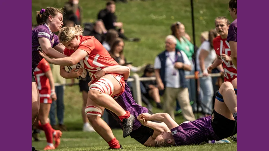 Two teams of female rugby players wearing purple tops, and red and white tops, playing against each other