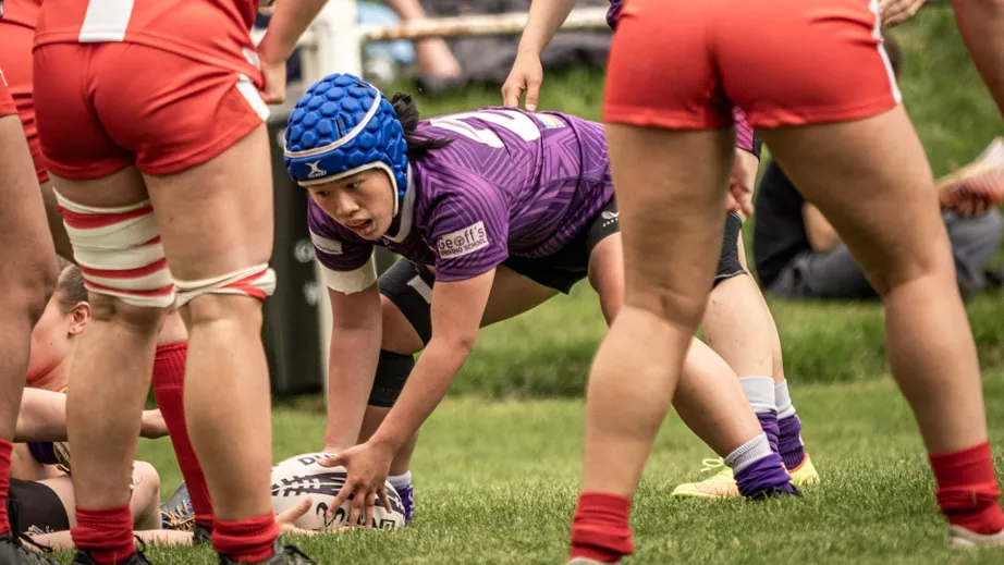 Two teams of female rugby players wearing purple tops, and red and white tops, playing against each other
