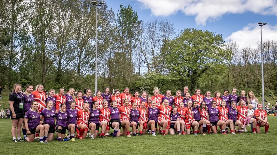 Two teams of female rugby players wearing purple tops, and red and white tops standing in a long line for the camera.