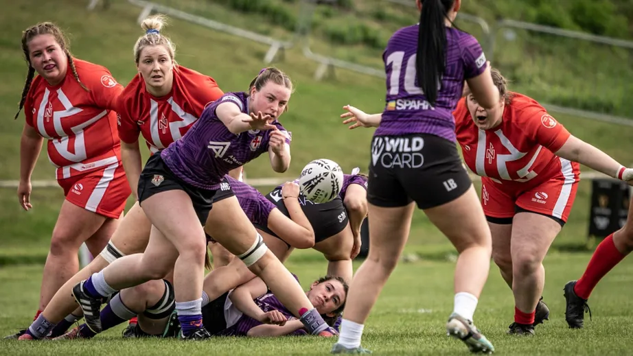 Two teams of female rugby players wearing purple tops, and red and white tops, playing against each other