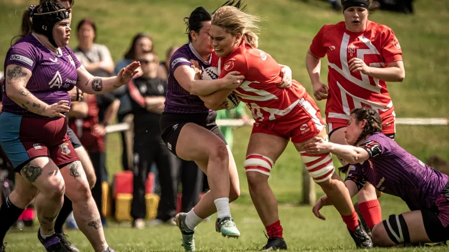 Two teams of female rugby players wearing purple tops, and red and white tops, playing against each other
