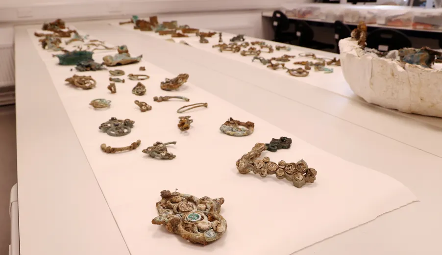 A variety of artefacts from the Melsonby Hoard laid out on a white table, including various pieces of corroded metalwork and a damaged cauldron.