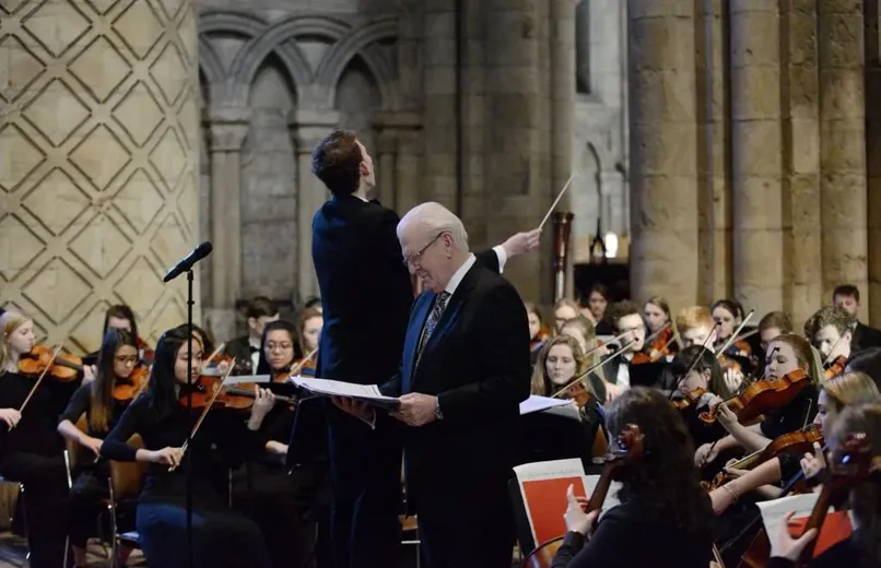 A white haired man looks down at a score, while a conductor behind him leads an orchestra