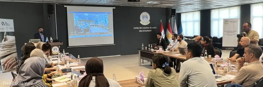A classroom with people sitting at desks looking at a screen while someone delivers a talk.