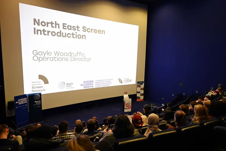 Woman standing speaking to tiered lecture theatre full of young adults, with large screen to her right