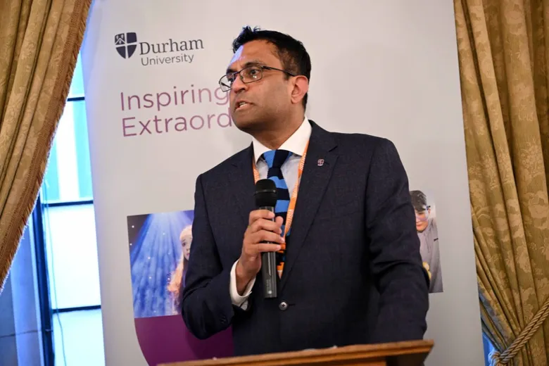 A man in a suit and tie speaking into a handheld microphone, standing at a lectern