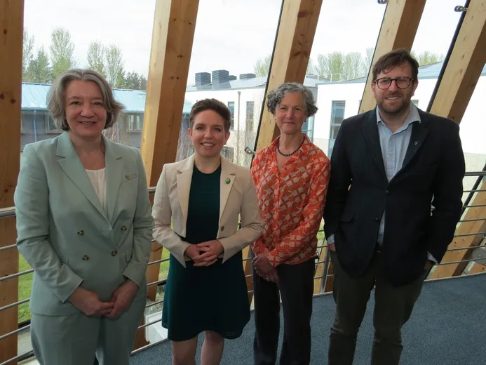 Four people looking at the camera, smiling. Behind them is a glass wall, through which can be seen other buildings and a grassed area