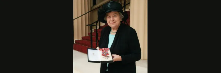 Professor Dame Rosemary Cramp in black hat and jacket holds her Damehood outside Buckingham Palace