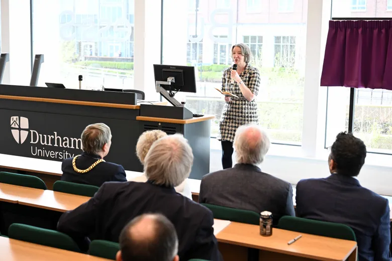 Woman holding microphone speaking to guests seated in tiers, backs of heads visible