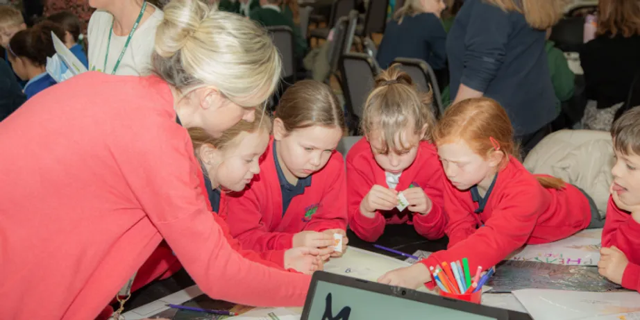 Schoolgirls in red jumpers and a teacher in a pink cardigan lean over work on a desk