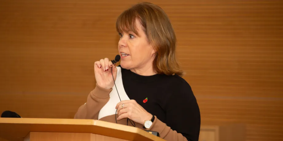 A woman with brown hair wearing a black and beige top speaking into a small microphone at a lecturn