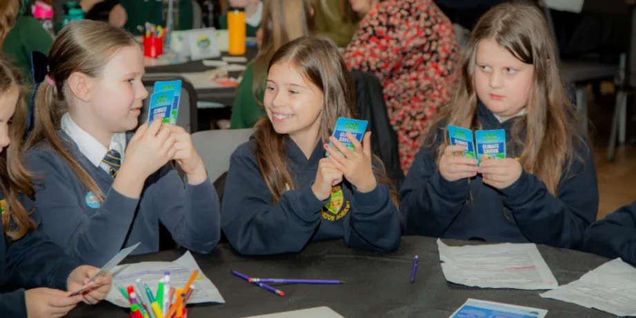 Three schoolgirls in blue uniforms are holding cards and smiling at each other