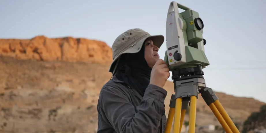 A dark-haired woman wearing a light-coloured, brimmed hat looks through camera-like equipment in a desert landscape