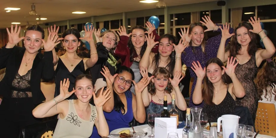 A group of 7 students in evening dress in a line at a round table with their hands next to their ears like antlers