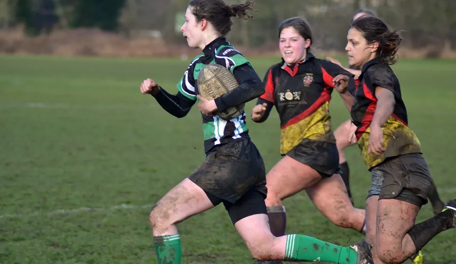 Cuth’s female rugby player in green kit running with a rugby ball and being chased by 2 opponents.