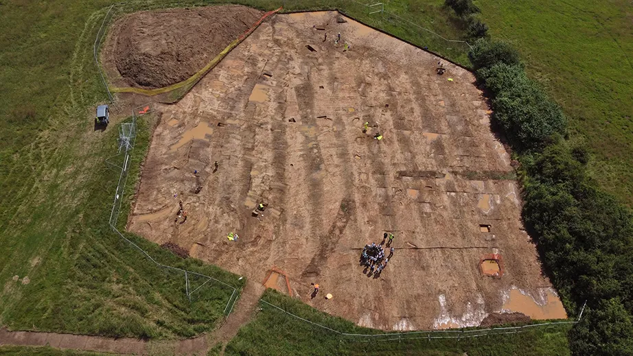 An open air excavation seen from above with features outlined in brown soil and a group of people in a circle as part of an open day