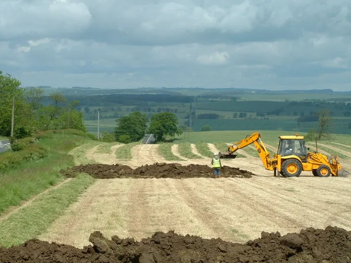 An archaeologist watching a yellow JCB excavate a trench in a field under a cloudy sky
