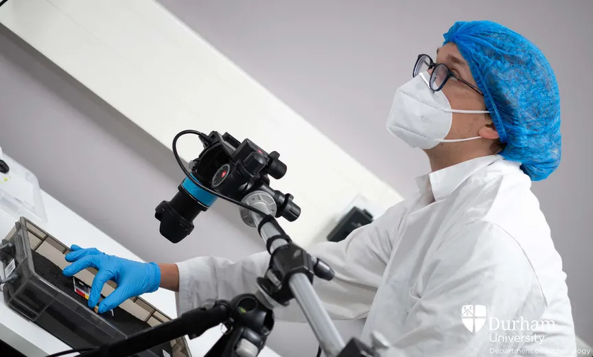 Student wearing Personal Protective Equipment uses a camera to scan a small object inside a foam-lined box. The student is looking intently at a screen that is out-of-frame.