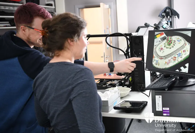 Two students stand inside the Digital Visualisation lab and look at a screen displaying a photo of a decorated object.