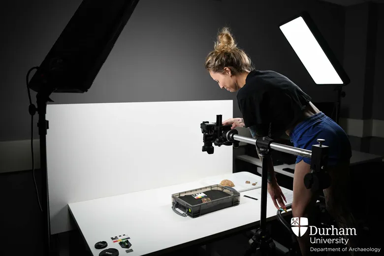 Student standing on stepladder above a well-lit worktop with an object inside a dark foam-lined tray. There is a camera attached to a tripod directly above the object.