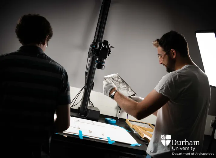 Two people inside a darkened Digital Visualisation Lab, one is wearing gloves and holding a piece of film or paper. The people stand next to a brightly lit worktop that has a camera above it and lamps on either side.