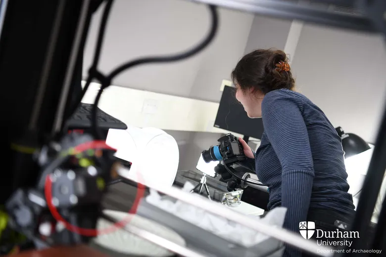 Student faces away from camera. She is smiling and holds a camera pointed towards an object that is not visible in this photo.