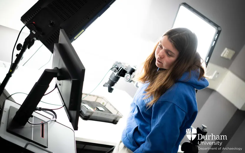 A student wears a blue Archaeology hoodie and looks at a screen that faces away from the viewer. She stands next to a brightly lit surface with a camera positioned above an object inside a tray.