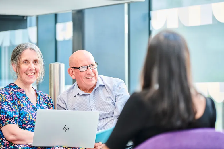 Tow members of staff, one male and one female, are chatting to a female student. The staff are facing towards camera, the student facing away.