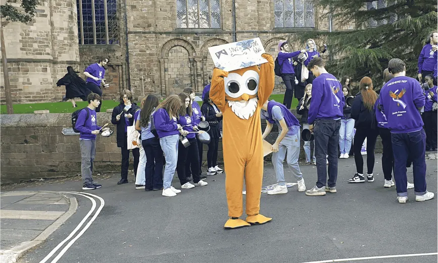 An owl mascot holding a banner with students with pots and pans in the background