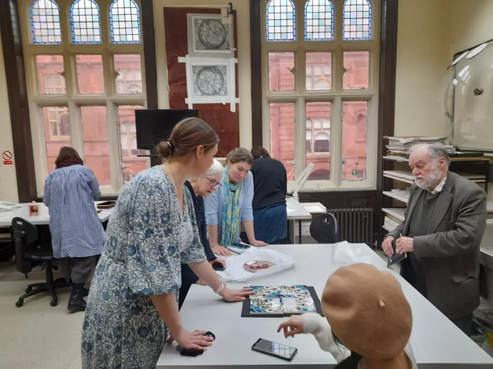 A group of people standing around a table examining tiles and a plate from the De Morgan foundation