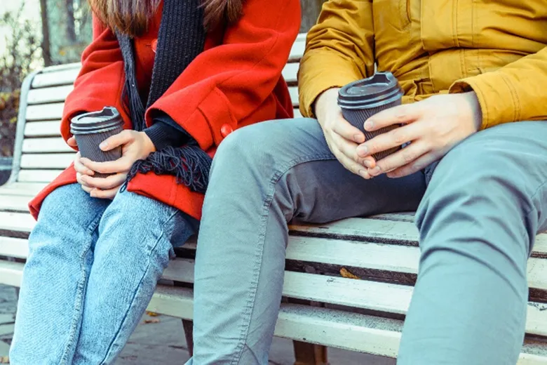 two students sitting on a bench holding cups of coffee