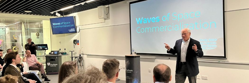 Dr Andrew Aldrin stands in front of a display screen and delivers a talk to a room full of people.