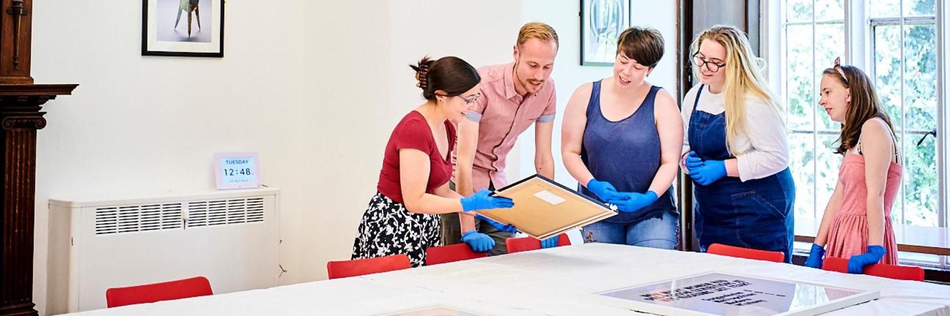 A group of people wearing gloves gather round as a woman shows them a framed picture.